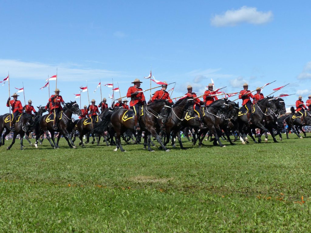 A group of Royal Canadian Mounted Police (RCMP) officers in red serge on horseback, riding across a grassy field with lances, during a ceremonial event.