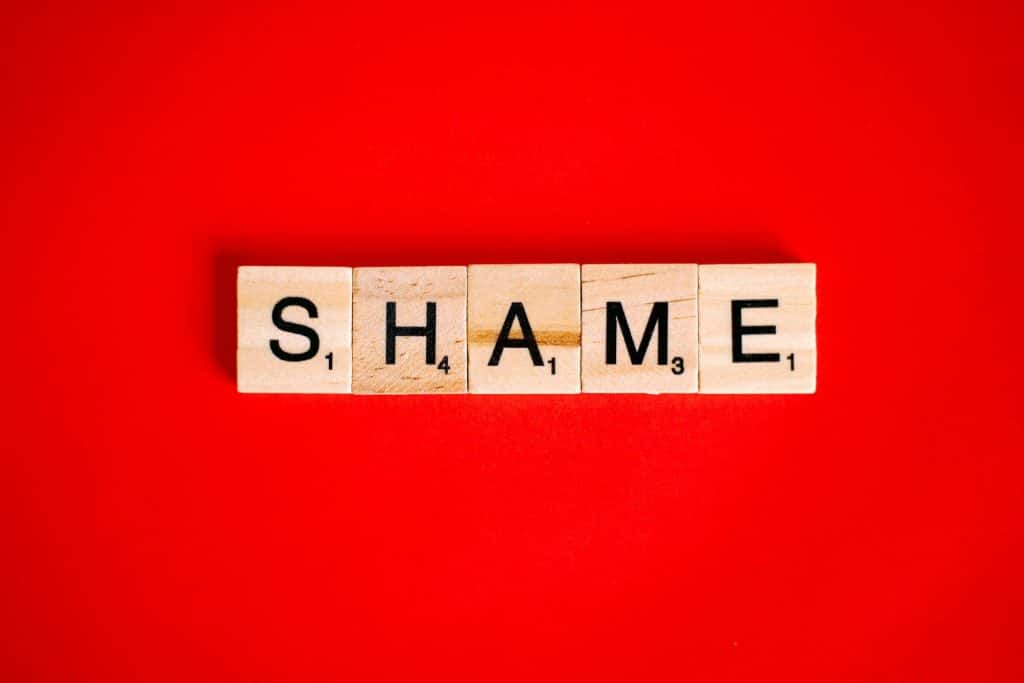 The word "SHAME" spelled out in wooden Scrabble-like tiles on a vibrant red background.