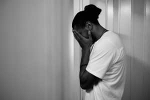 A black and white photo of a man standing alone, with his face in his hands. He appears to be feeling shame, sad or is in distress.