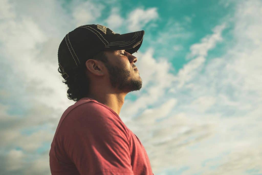 A man with a beard and baseball cap, looking up with his eyes closed and a calm expression on his face against a backdrop of a clear blue sky and clouds.