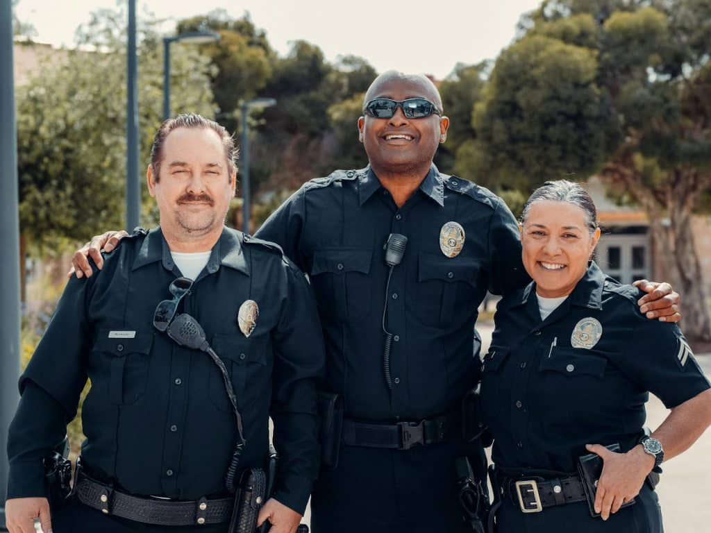 A diverse group of three smiling police officers, two men and one woman, standing outside in uniform with their arms around each other.