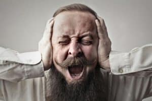 A man with a beard and mustache screaming, with his eyes squeezed shut and hands covering his ears, a visual representation of intense emotional dysregulation.