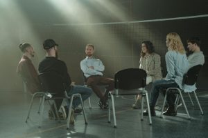 A group of six people in a dimly lit room, sitting in a circle on folding chairs, with a volleyball net in the background, appearing to be in a support group session.